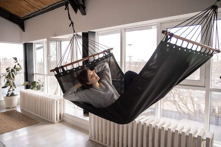 Young female in casual wear lying in comfortable hammock with eyes closed leaned on hands while sleeping in apartment near window and radiators with potted plant